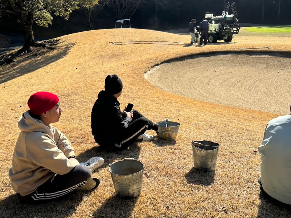 Workers seated beside a golf course bunker, observing maintenance activities in progress.