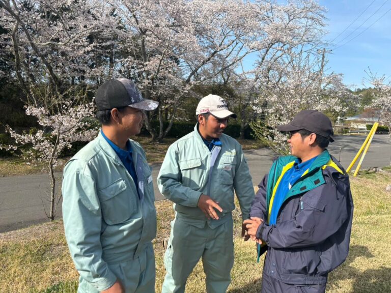 桜の咲くゴルフ場で、作業服を着た3人の男性が屋外で会話をしている様子。